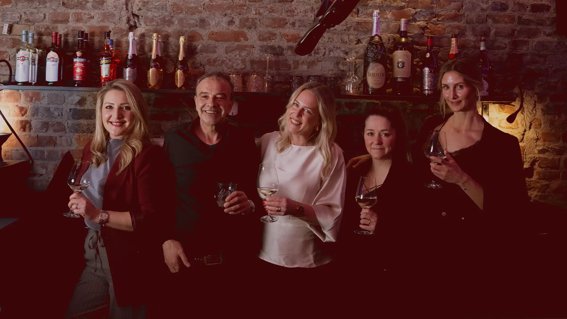 Giovanni und Gäste stehen mit gefüllten Weingläsern vor einer Backsteinwand